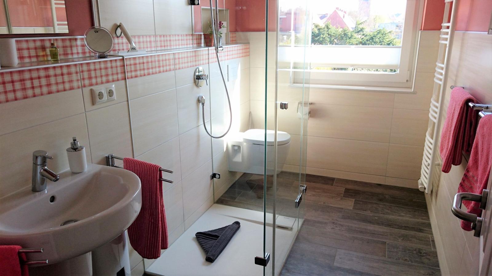 Badezimmer mit Dusche, Waschbecken und Toilette. Rote Handtücher und Fenster mit Blick.