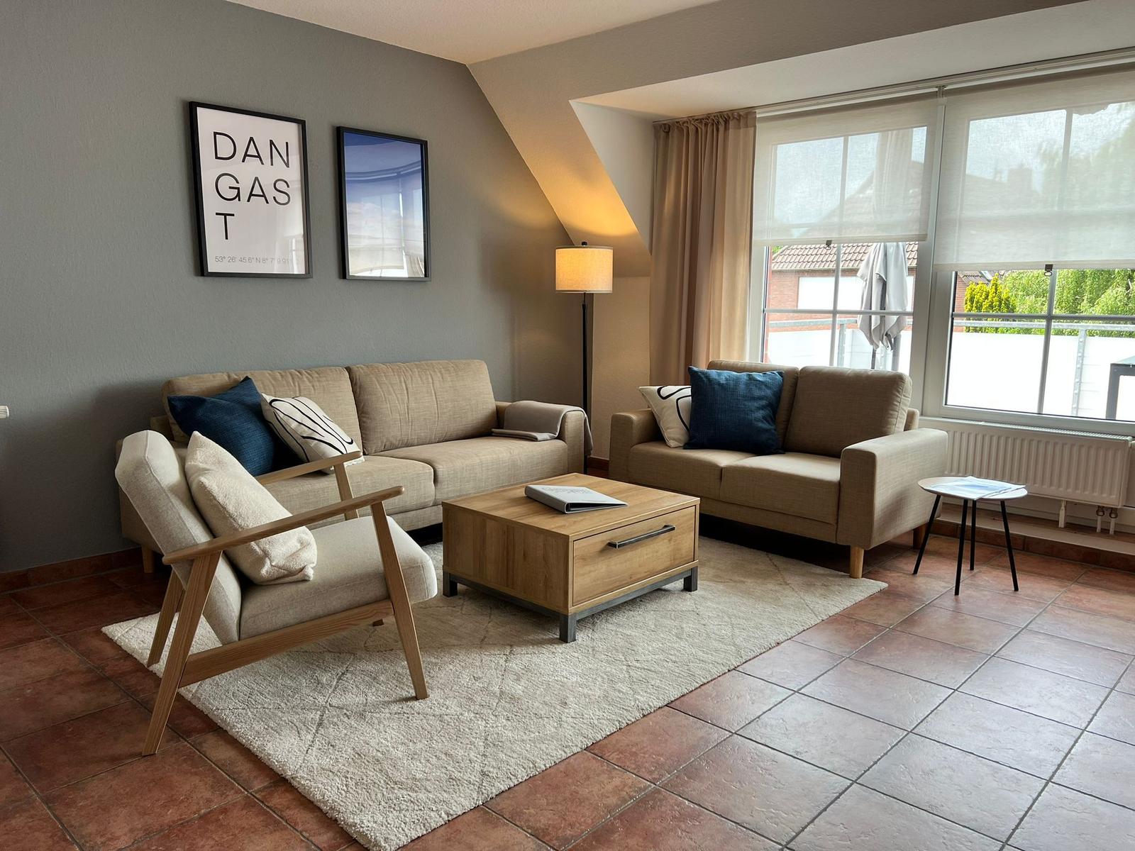 Wohnzimmer mit Sofa, Sessel, Couchtisch und Fenster. Wandbilder und Teppich sind sichtbar.