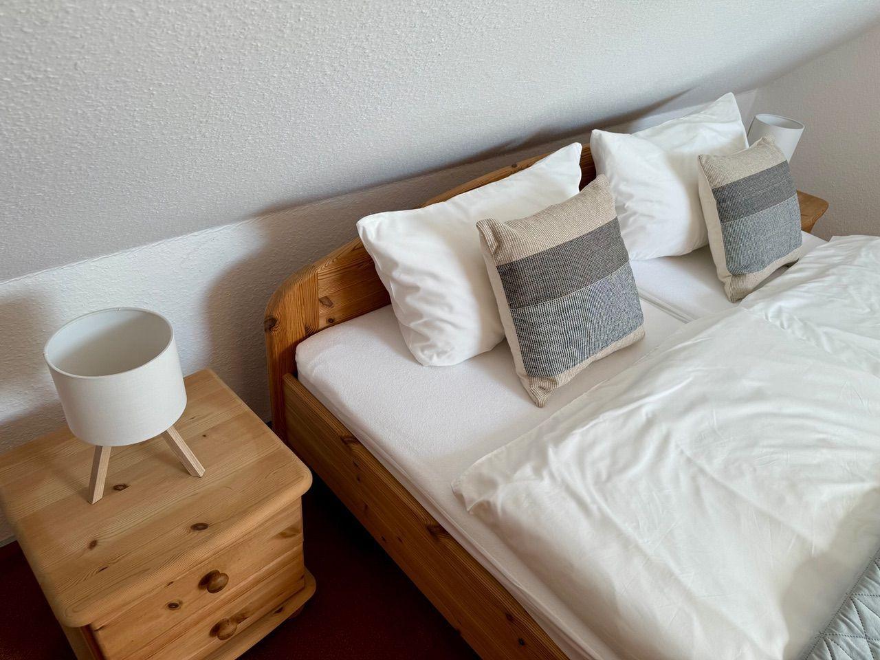 Ein Bett mit weißen Laken und Kissen, daneben ein Nachttisch mit Lampe.