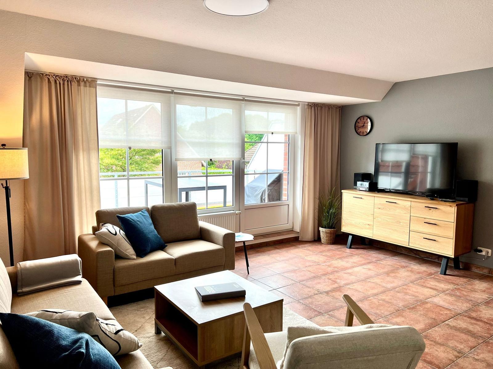 Wohnzimmer mit Sofa, Couchtisch und Fernseher. Fenster mit Blick auf Balkon.