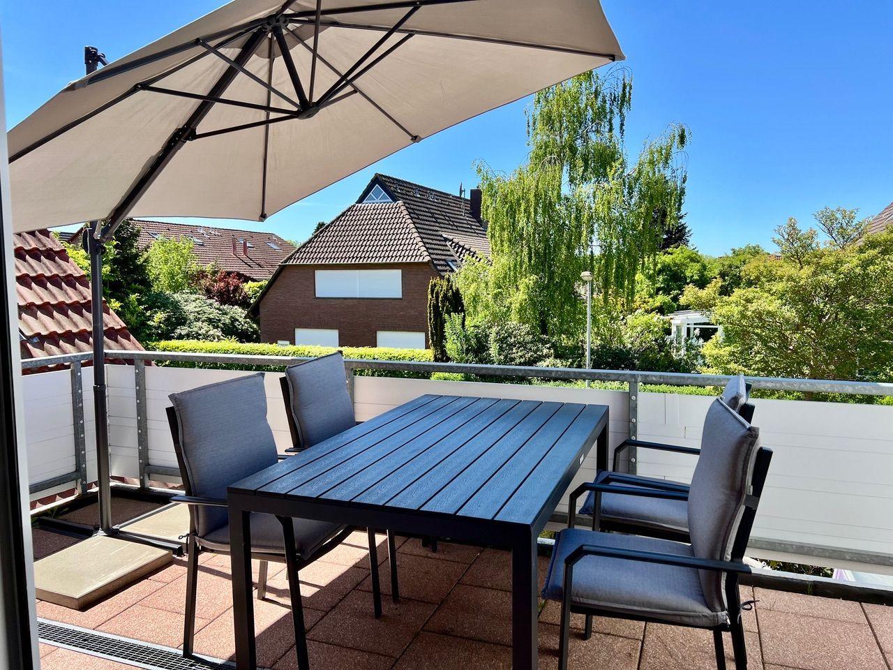 Terrasse mit Tisch, vier Stühlen und Sonnenschirm. Blick auf begrünte Umgebung und Nachbarhäuser.