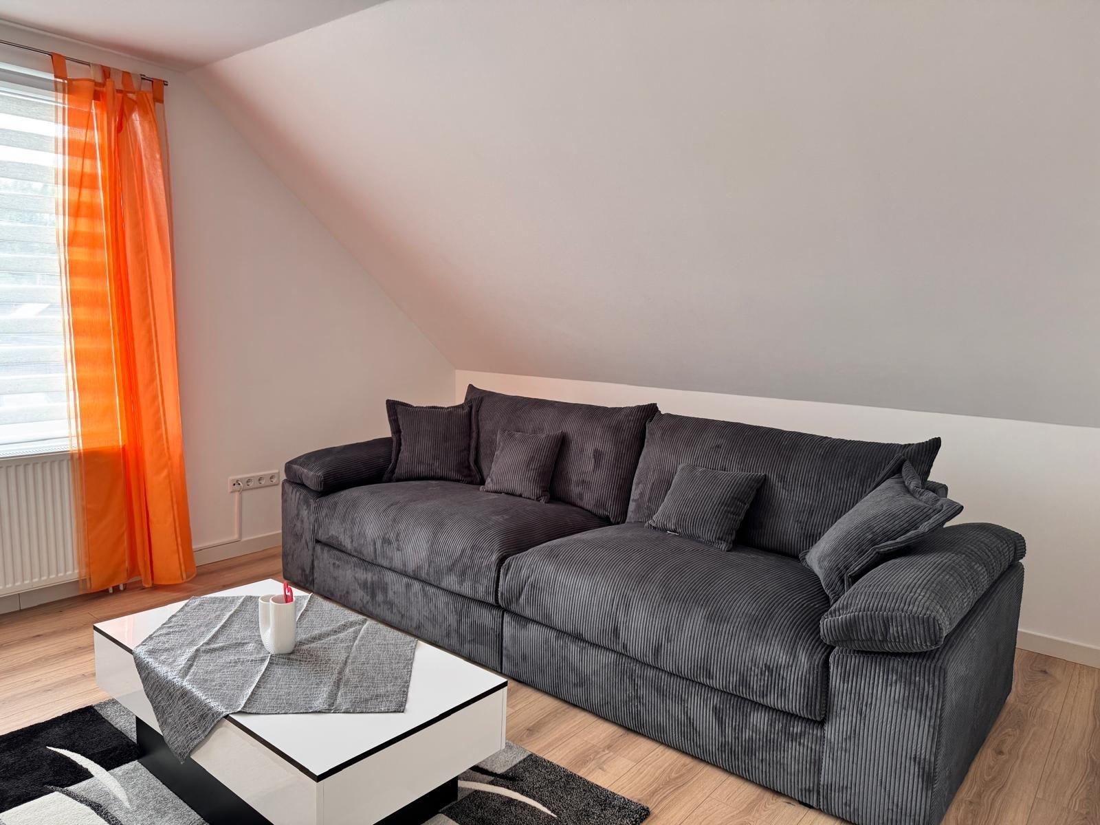 Ein Wohnzimmer mit grauem Sofa, weißem Tisch und orangefarbenen Vorhängen.