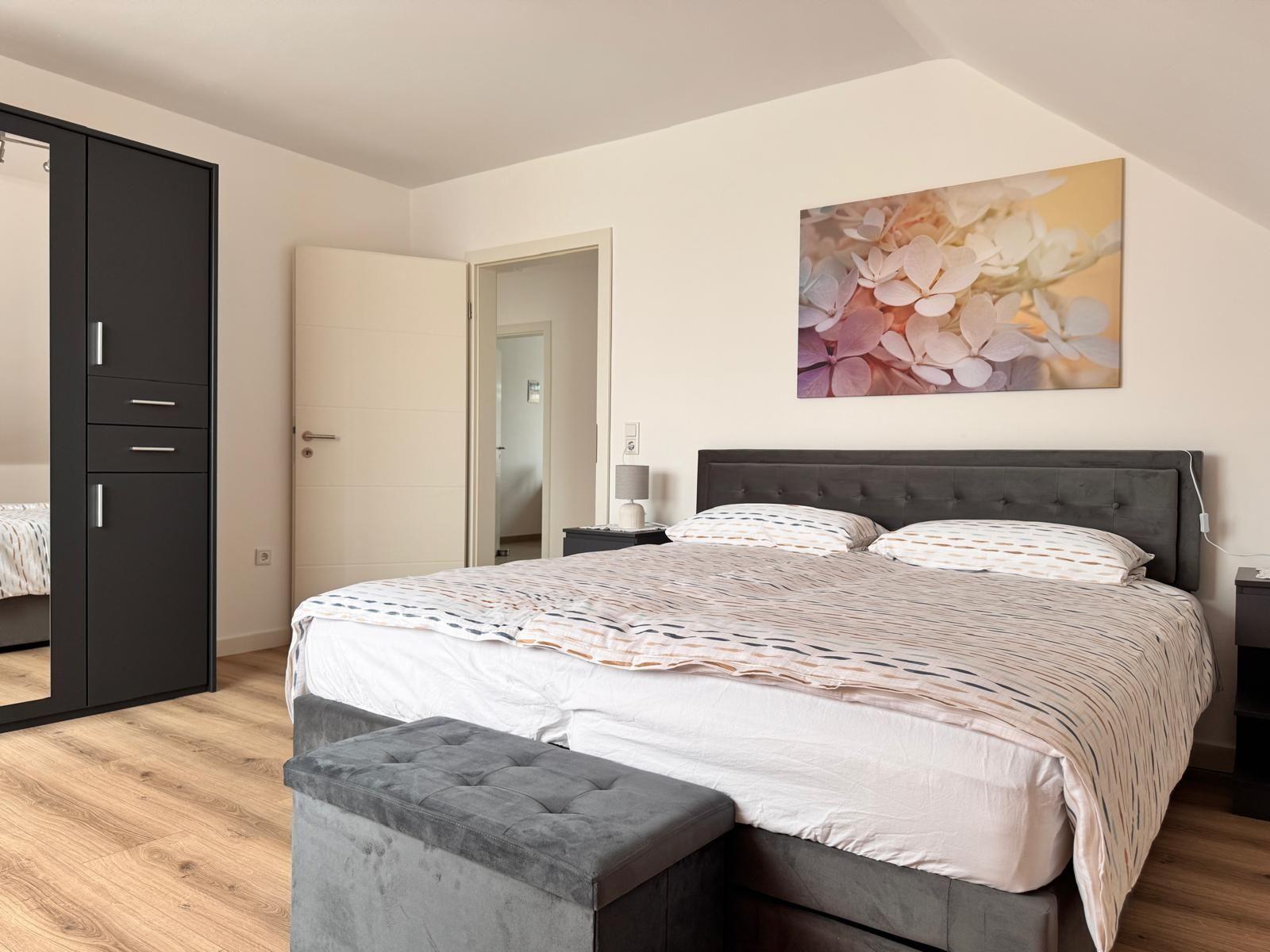 Schlafzimmer mit Doppelbett, Kleiderschrank und Wandbild mit Blumenmotiv.
