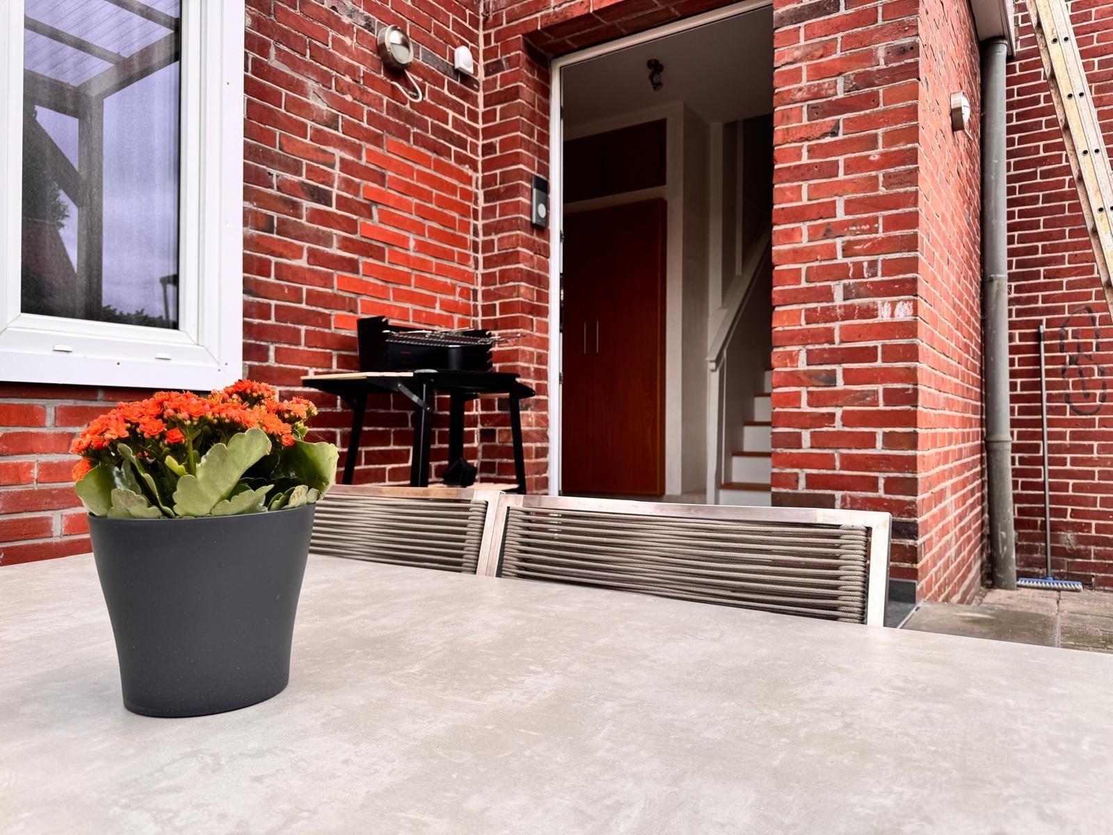 Roter Backsteinbau mit Terrasse, Grill und Pflanze. Eingang und Treppe sichtbar.