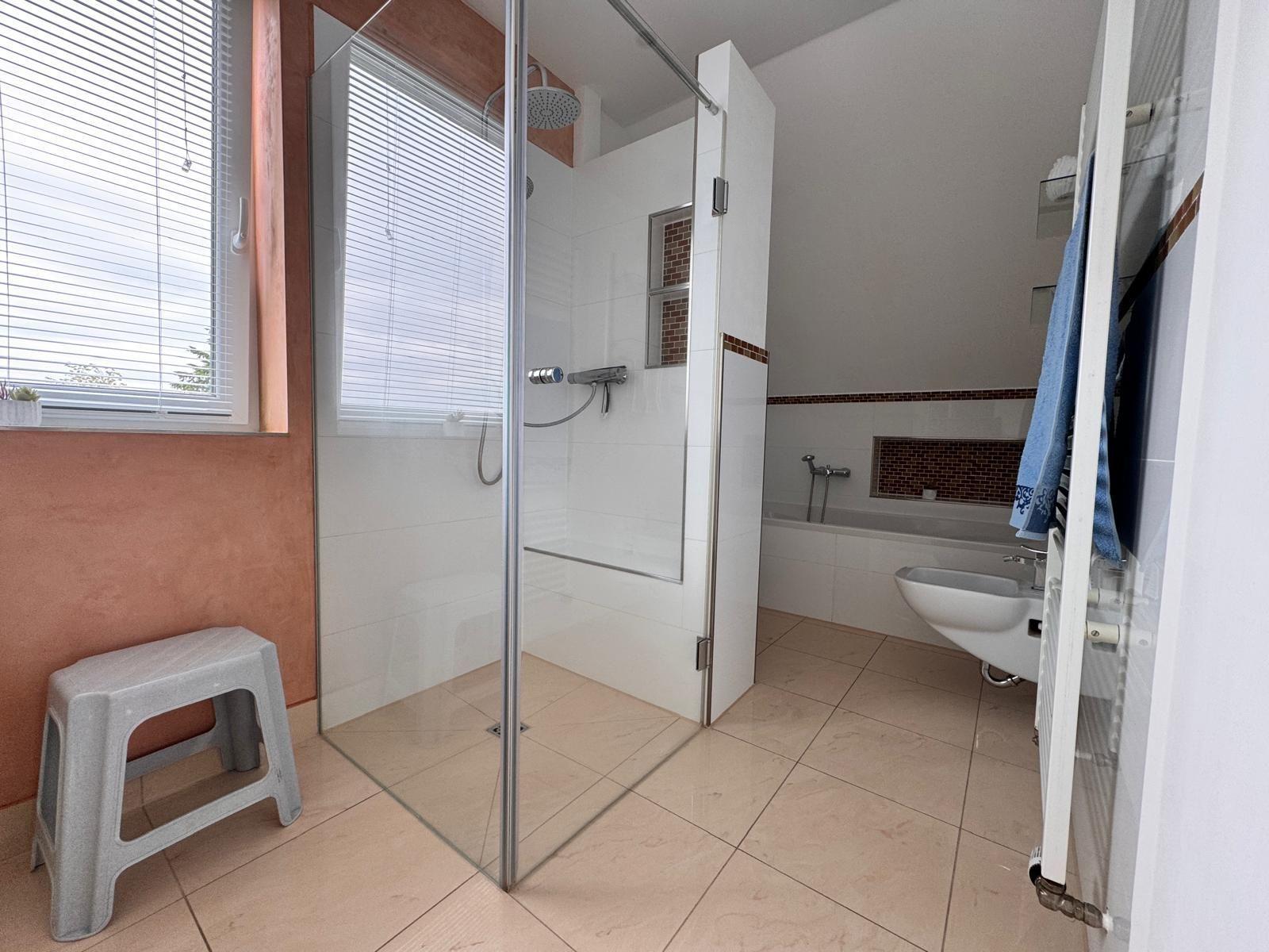 Badezimmer mit Dusche, Badewanne und Toilette. Fenster mit Jalousie und grauer Hocker.