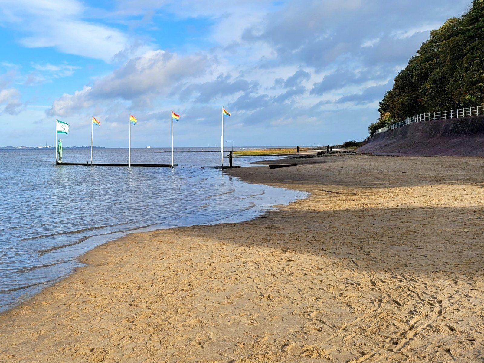 Strand mit Steg und Flaggen im Wasser, Himmel mit Wolken, Bäume am Ufer.