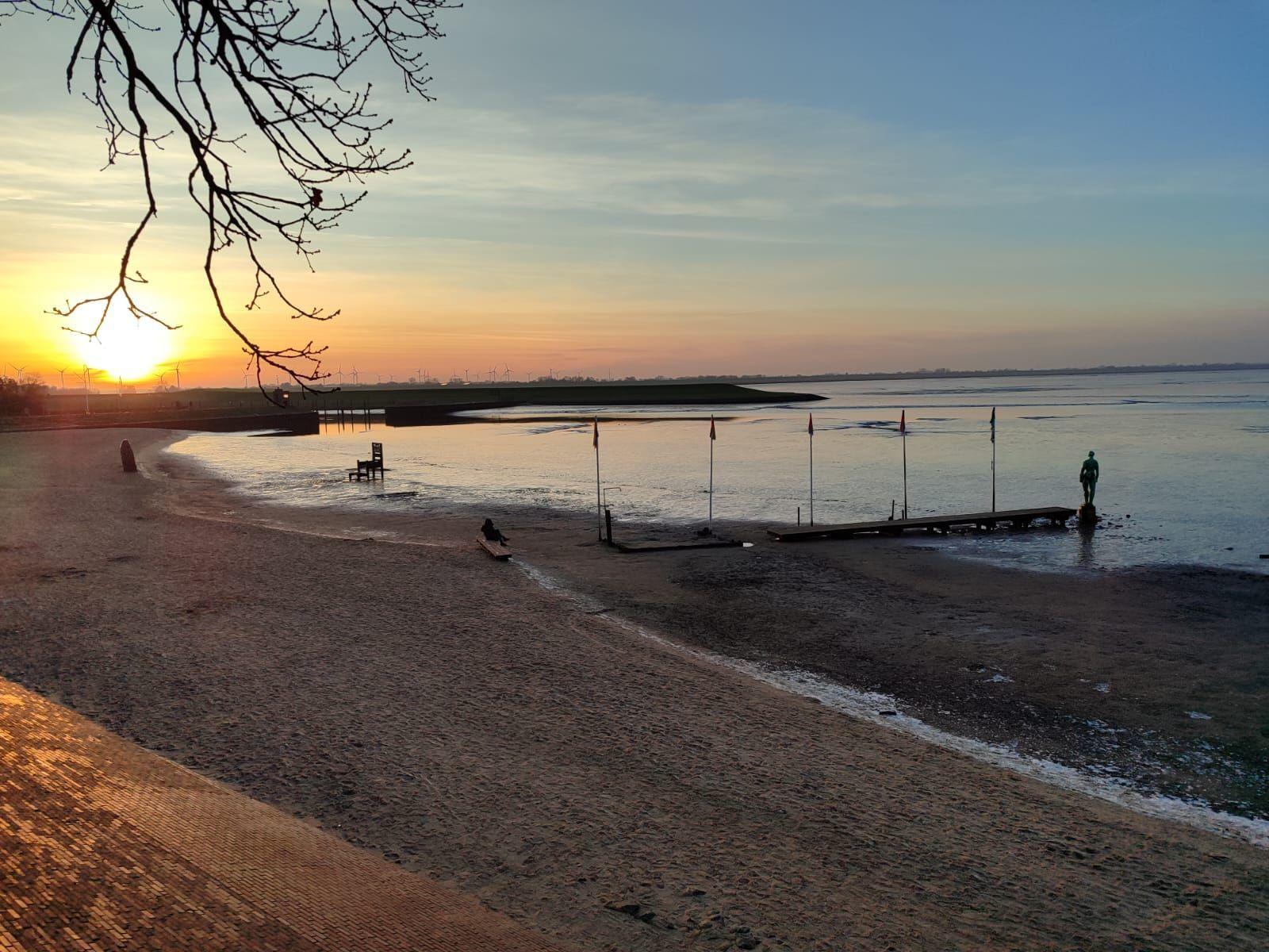 Sonnenuntergang am Strand mit Steg und Windkraftanlagen im Hintergrund.
