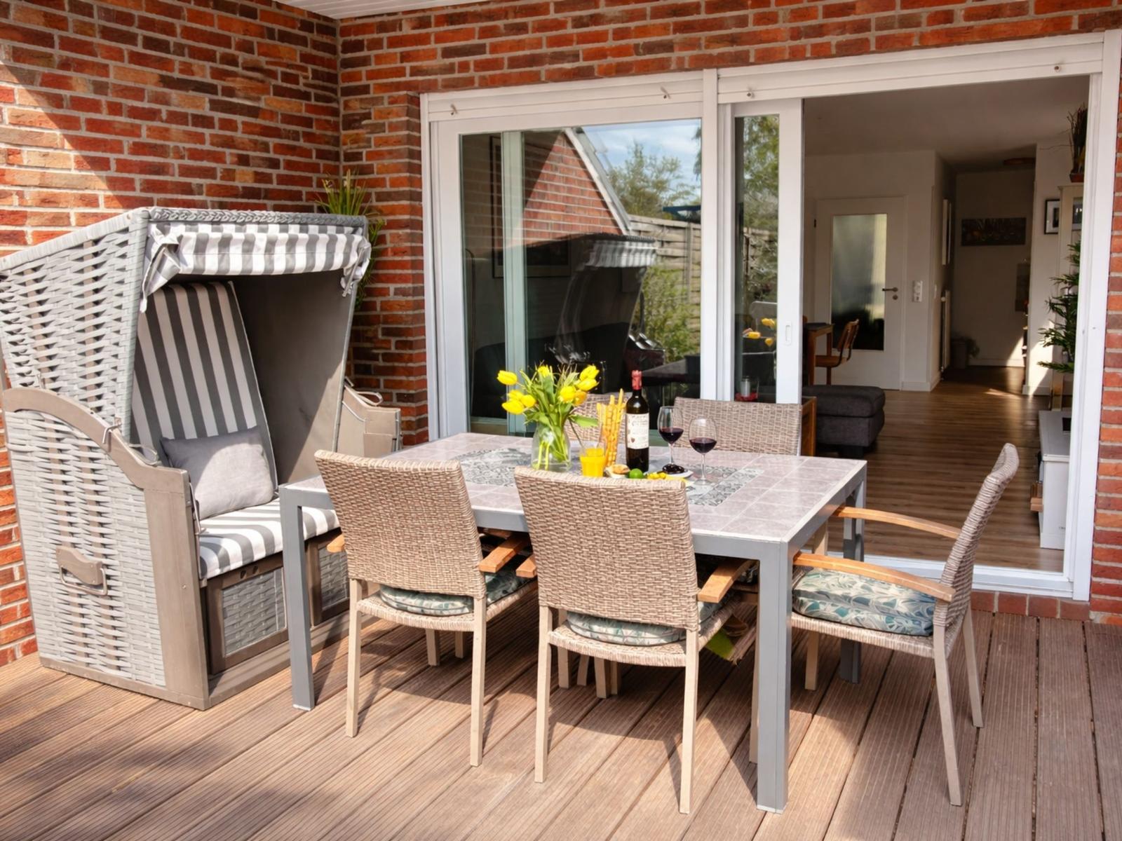 Terrasse mit Esstisch, Stühlen und Strandkorb. Durchgang ins Haus. Holzdecke und Backsteinwand.