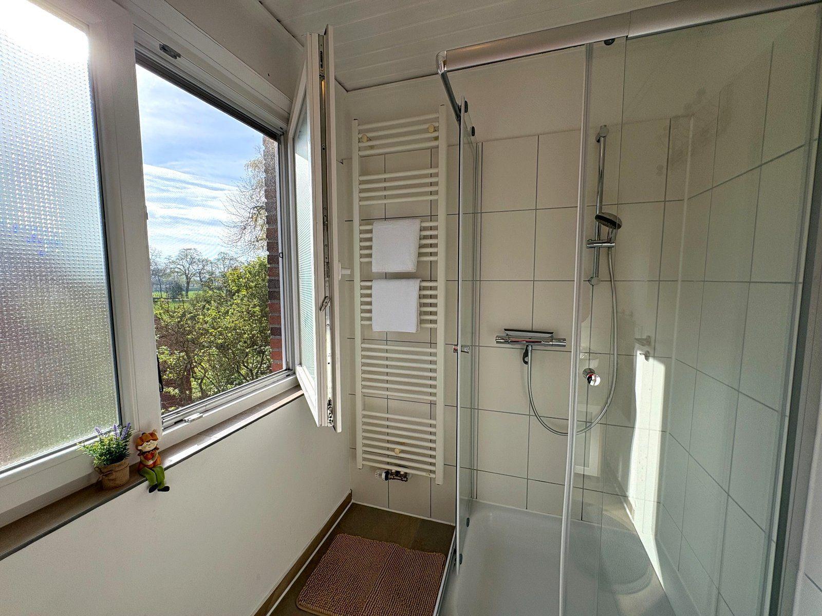 Badezimmer mit Dusche, Heizkörper und Fenster mit Blick auf Grünflächen.