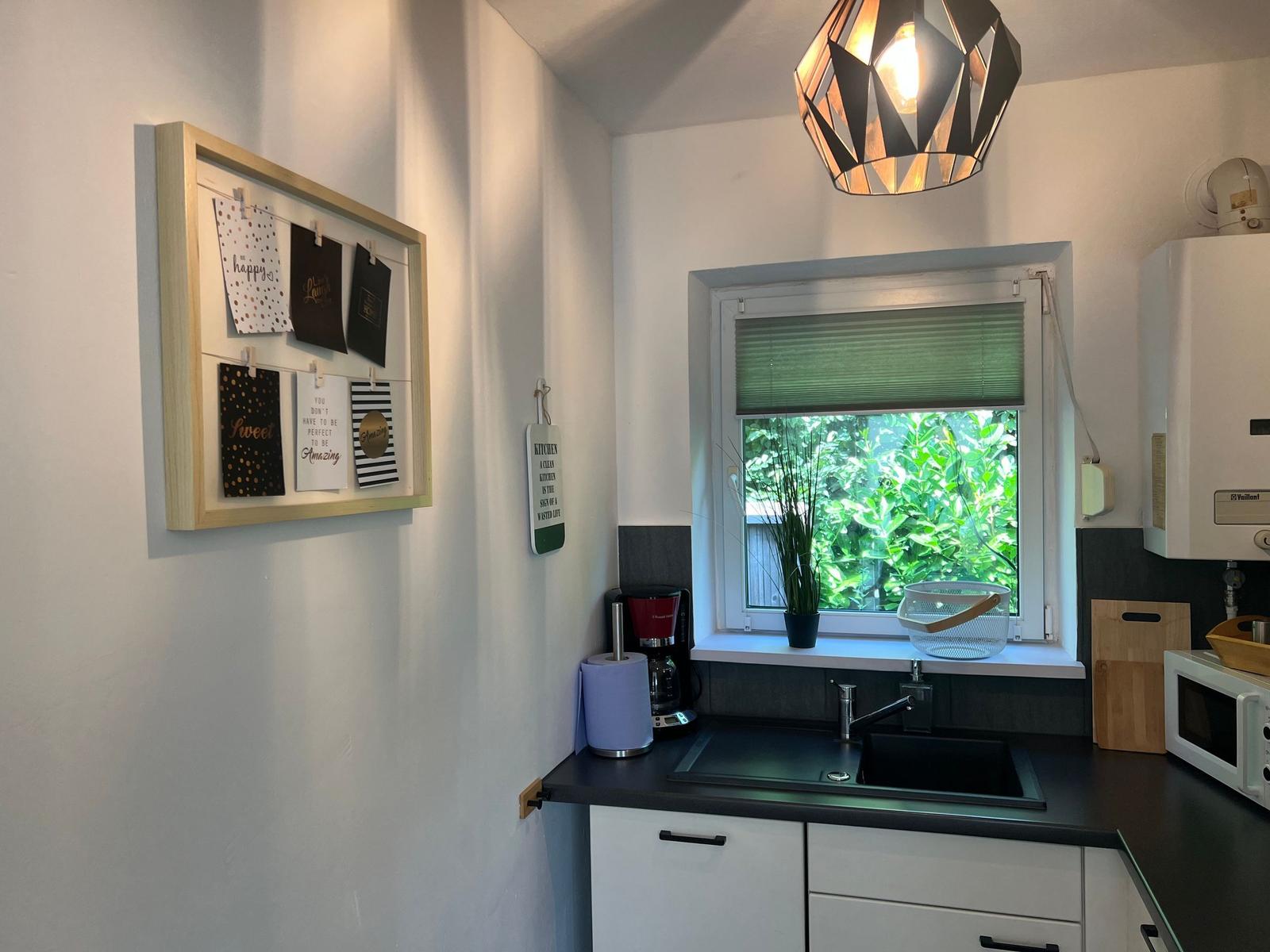 Küche mit Fenster, Spüle und Küchenzeile. Wand mit Bilderrahmen und Hängeschrank.