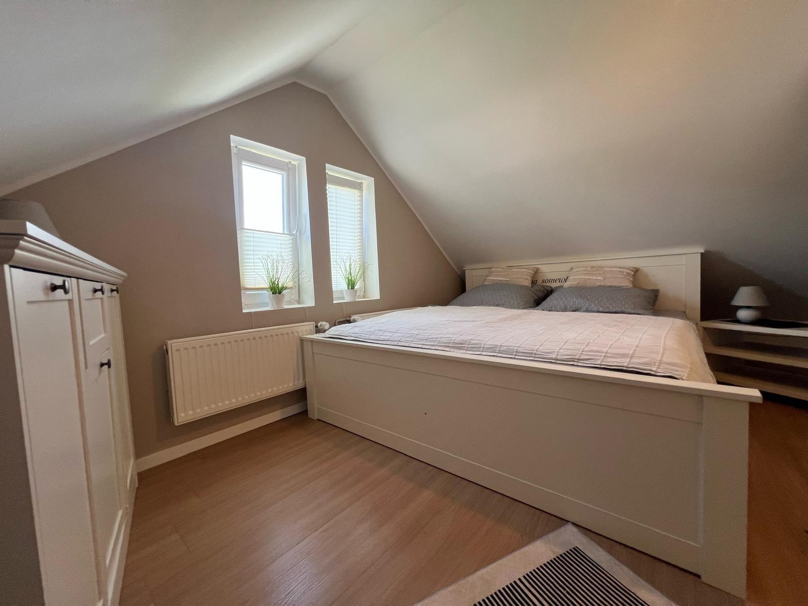 Schlafzimmer mit Dachschrägen, weißem Bett und zwei Fenstern mit Jalousien.