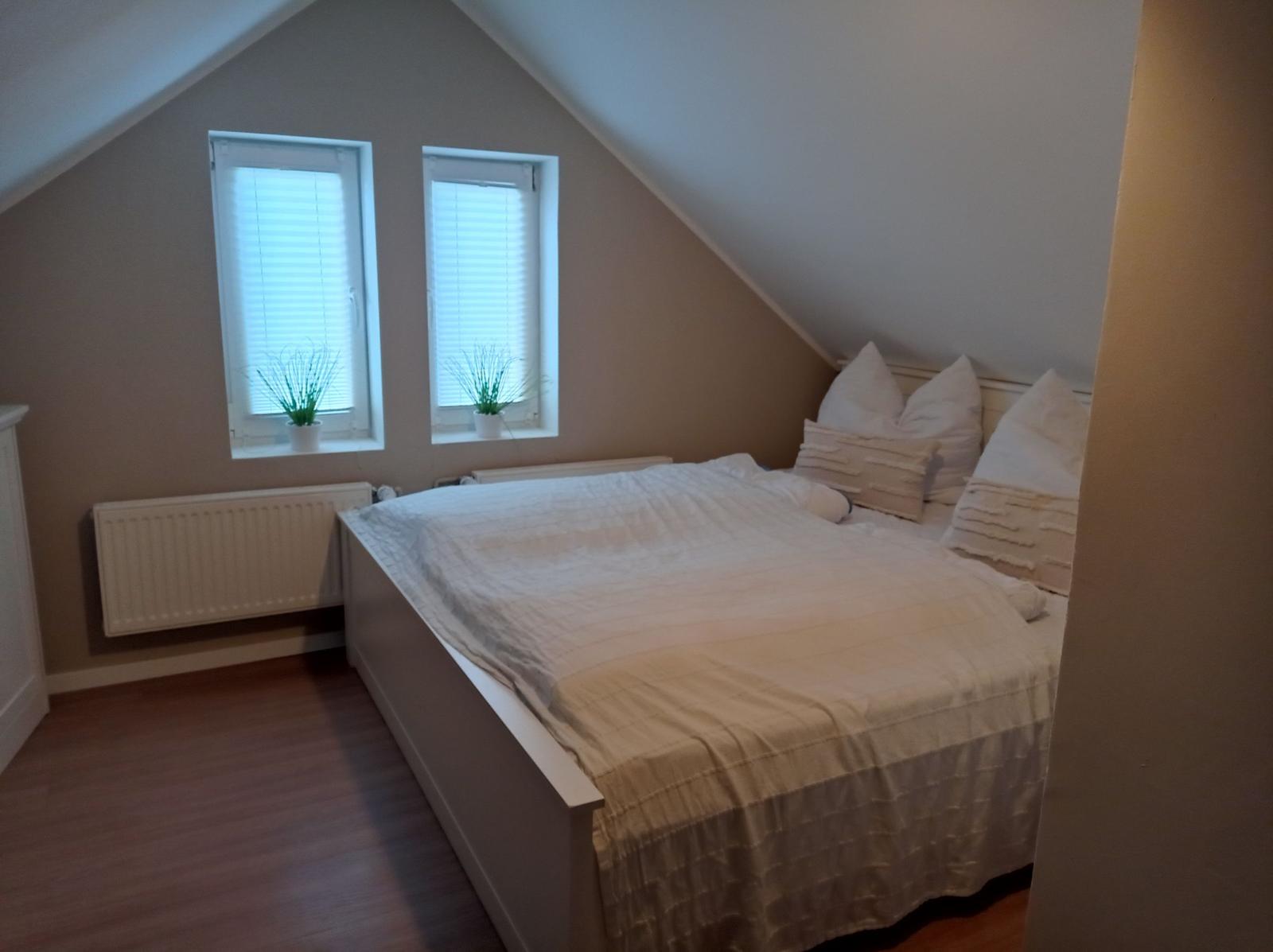 Schlafzimmer mit Dachschrägen, zwei Fenstern und Bett mit weißen Kissen.