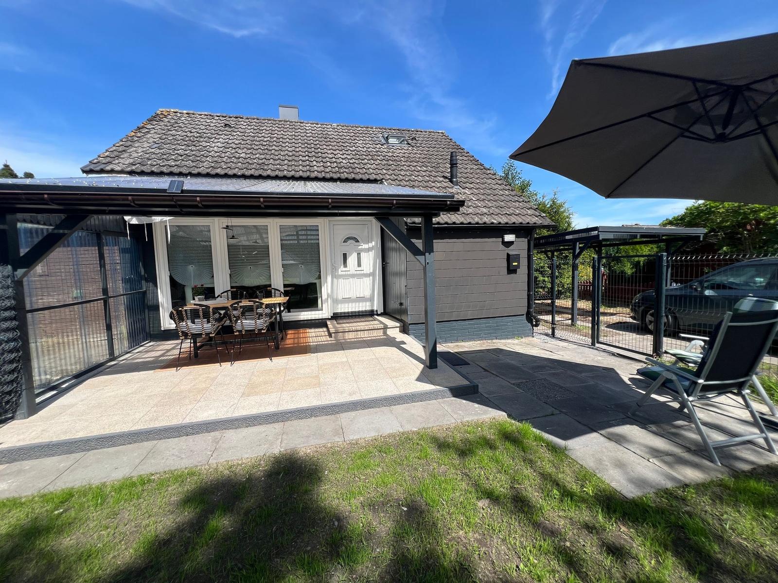 Haus mit Terrasse, Sitzgelegenheiten und Sonnenschutz. Grünflächen und gepflasterter Bereich.