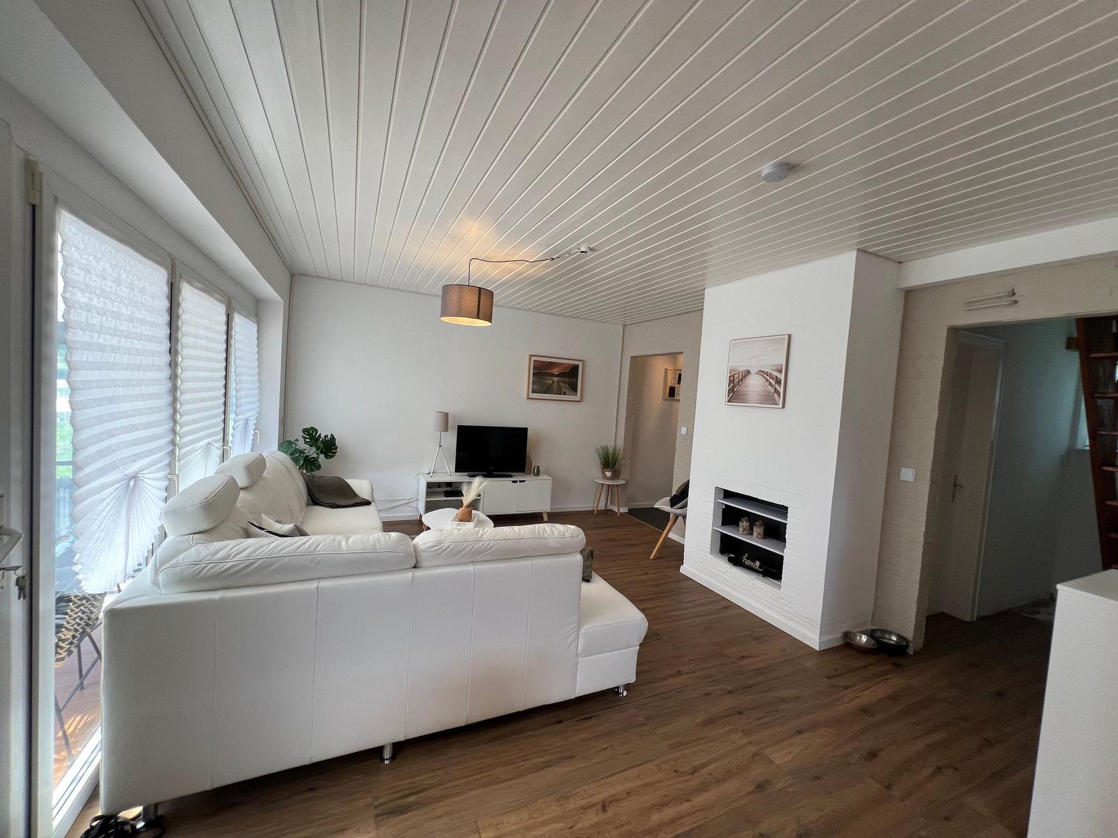 Wohnzimmer mit weißem Sofa, Holzboden und Kamin. Fenster mit Jalousien.