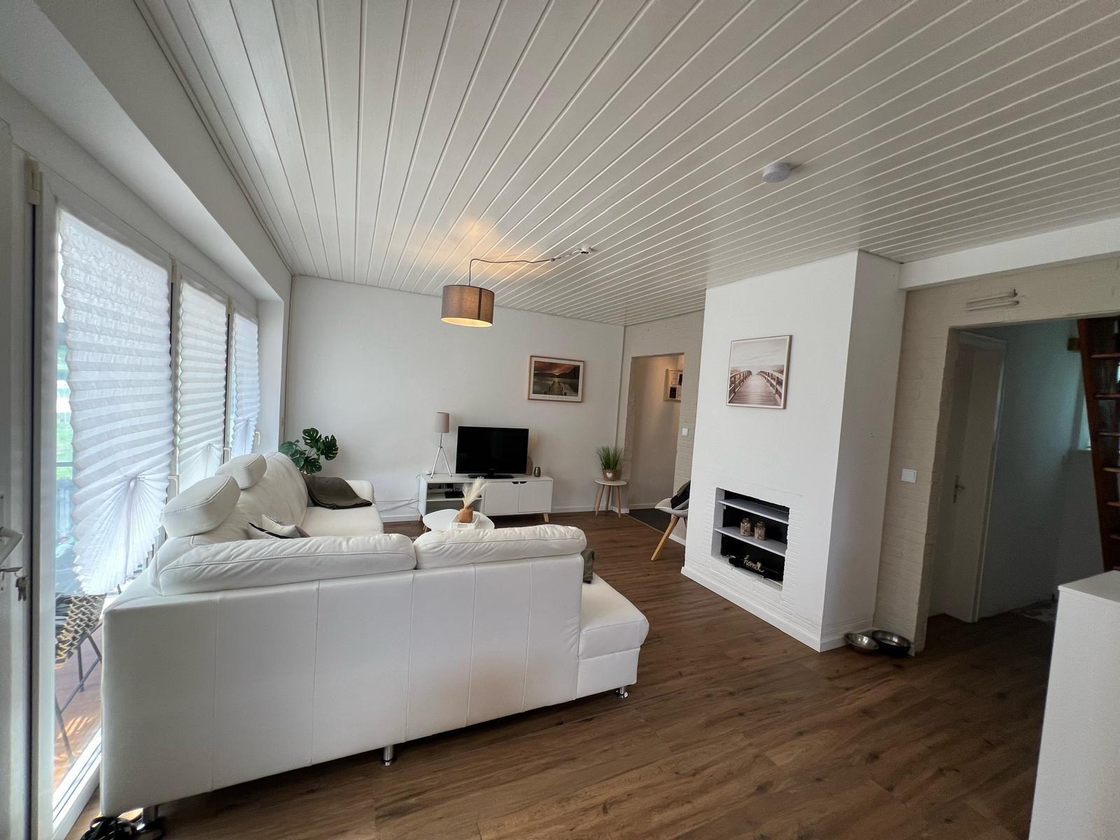 Wohnzimmer mit weißem Sofa, Holzboden und Kamin. Fenster mit Jalousien und TV.