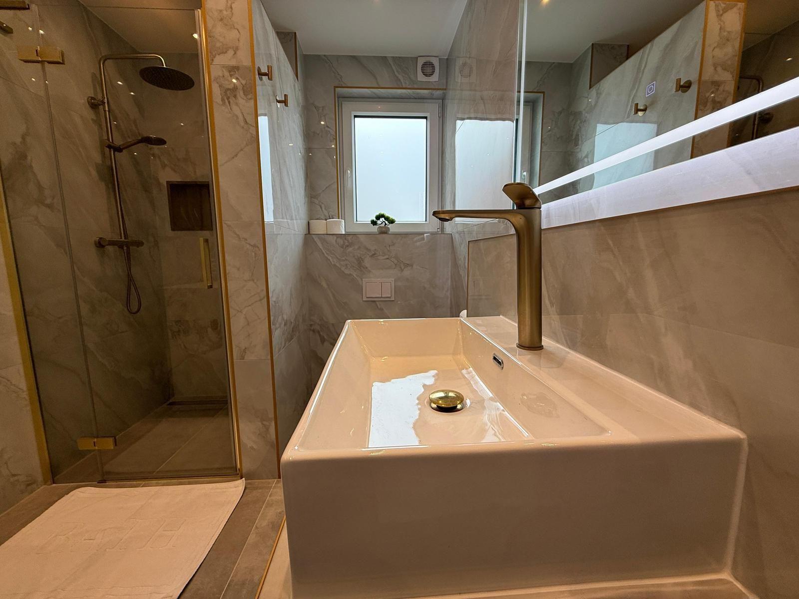 Bad mit Dusche, Waschbecken und Spiegel. Marmorwand und goldfarbene Armaturen.
