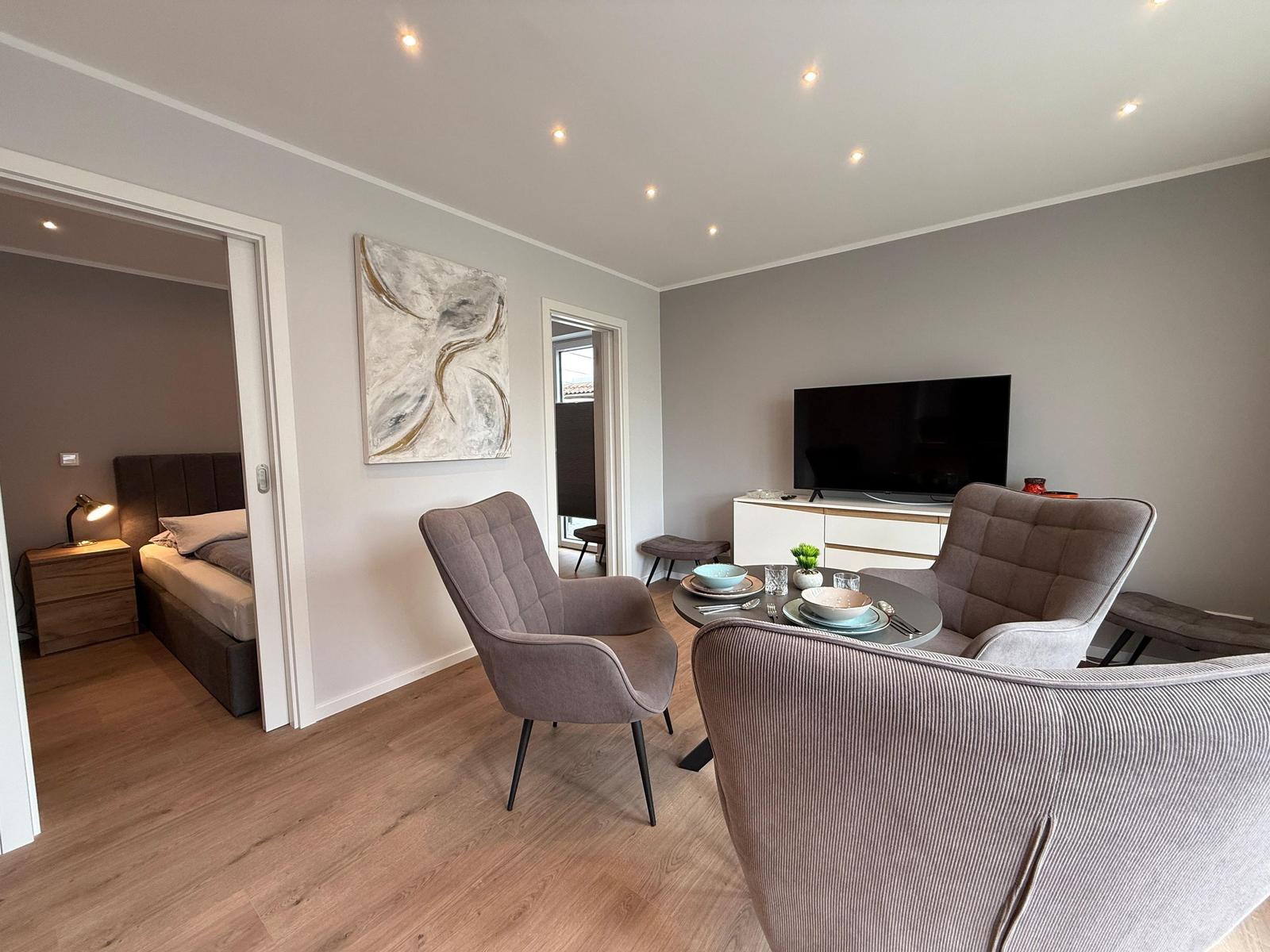 Wohnzimmer mit Sitzgelegenheiten, Fernseher und Blick in Schlafzimmer.