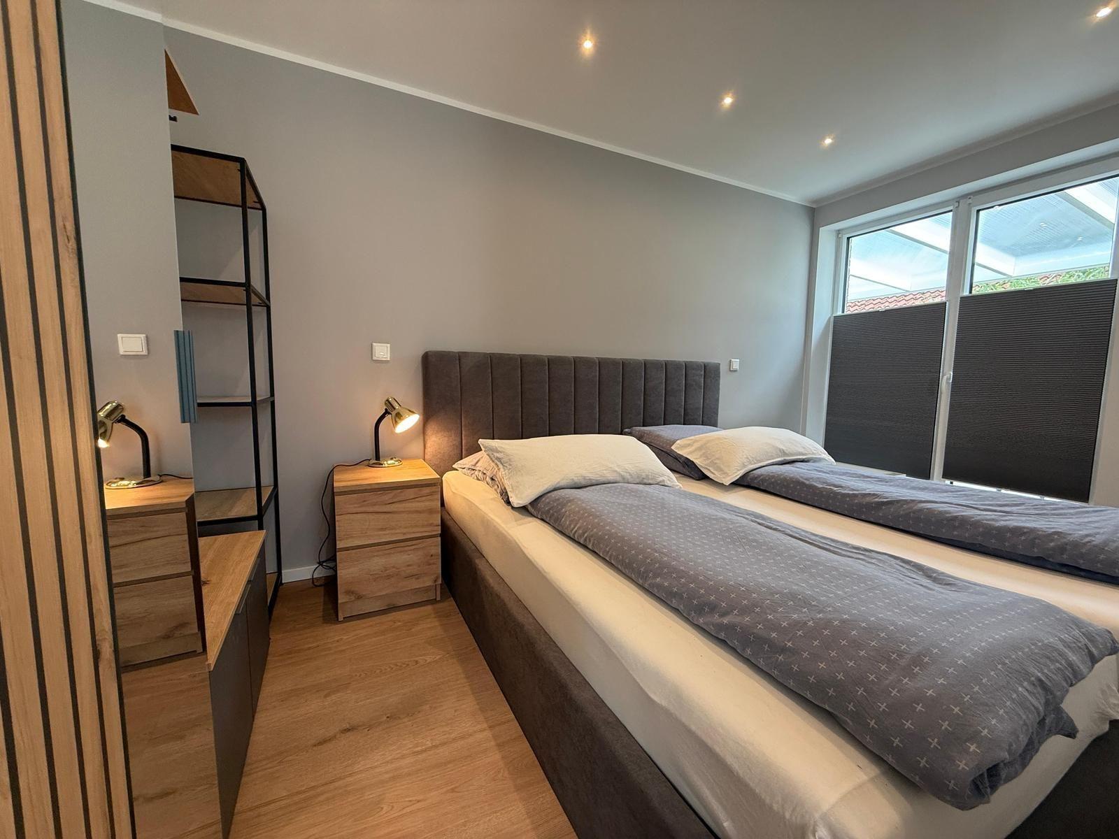 Schlafzimmer mit Doppelbett, Nachttisch und Schreibtisch. Fenster mit Jalousie.