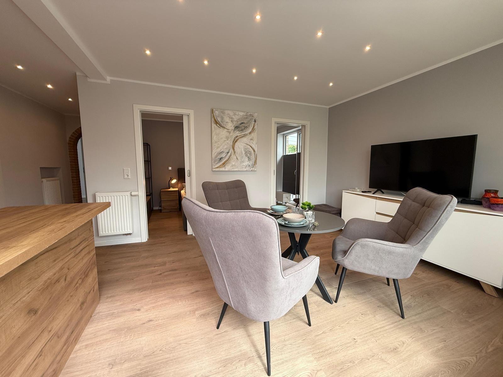 Wohn- und Essbereich mit grauen Sesseln, runder Tisch und Fernseher an Wand.