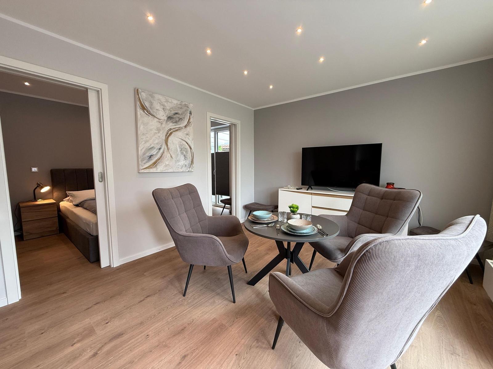 Wohn- und Essbereich mit drei Sesseln, runder Tisch und Fernseher. Schlafzimmer ist sichtbar.