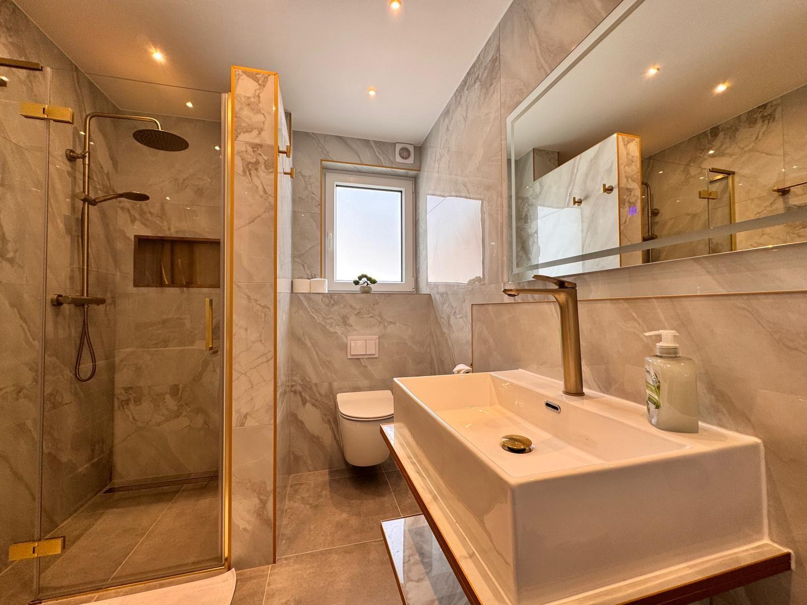 Badezimmer mit Dusche, Waschbecken und Toilette. Marmorwand und goldfarbene Details.