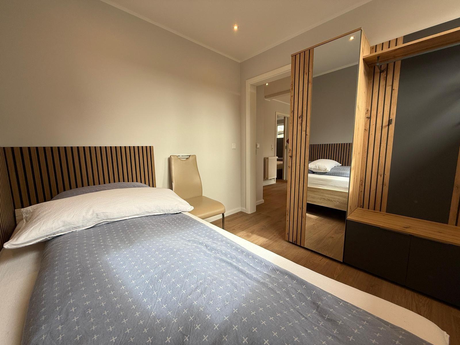 Schlafzimmer mit Bett, Stuhl und Spiegelschrank. Holzboden und Wandspiegel sichtbar.