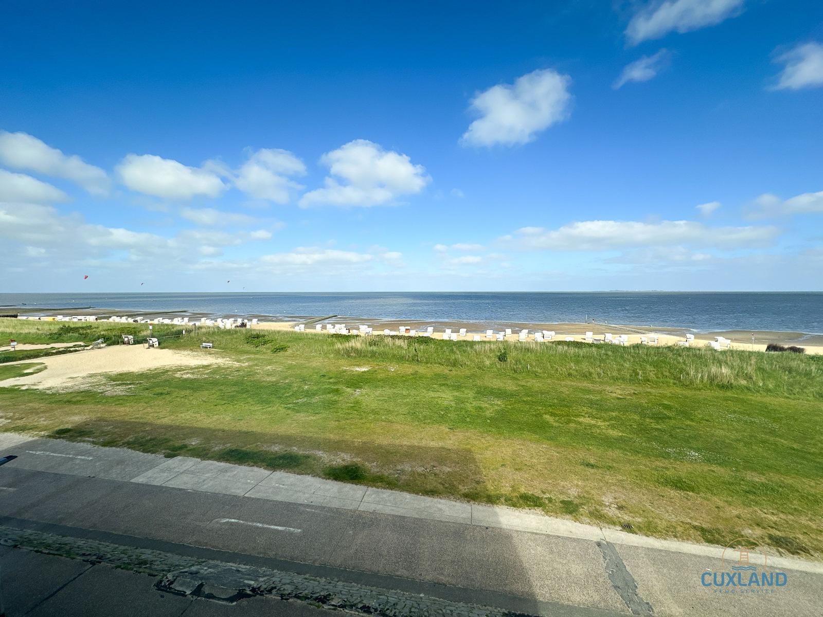 Weitläufiger Blick auf Strand mit weißen Liegestühlen und grüner Wiese unter blauem Himmel.