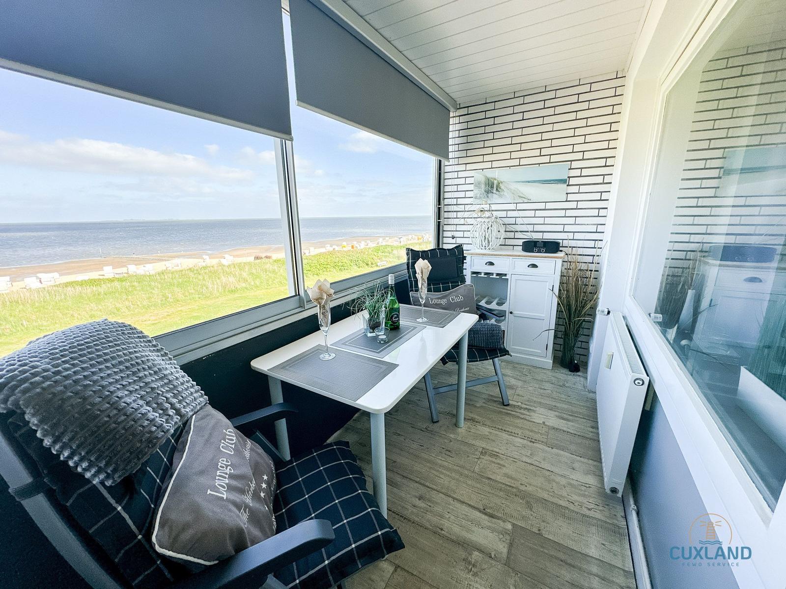 Terrasse mit Tisch, Stühlen und Meerblick. Weiße Wand mit Ziegeloptik und Fensterfront.
