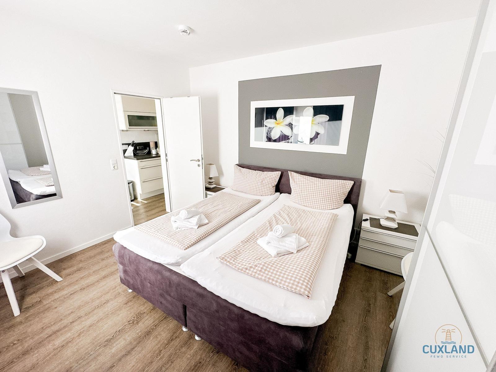 Schlafzimmer mit Doppelbett, Nachttisch und Bild an der Wand. Küche im Hintergrund.