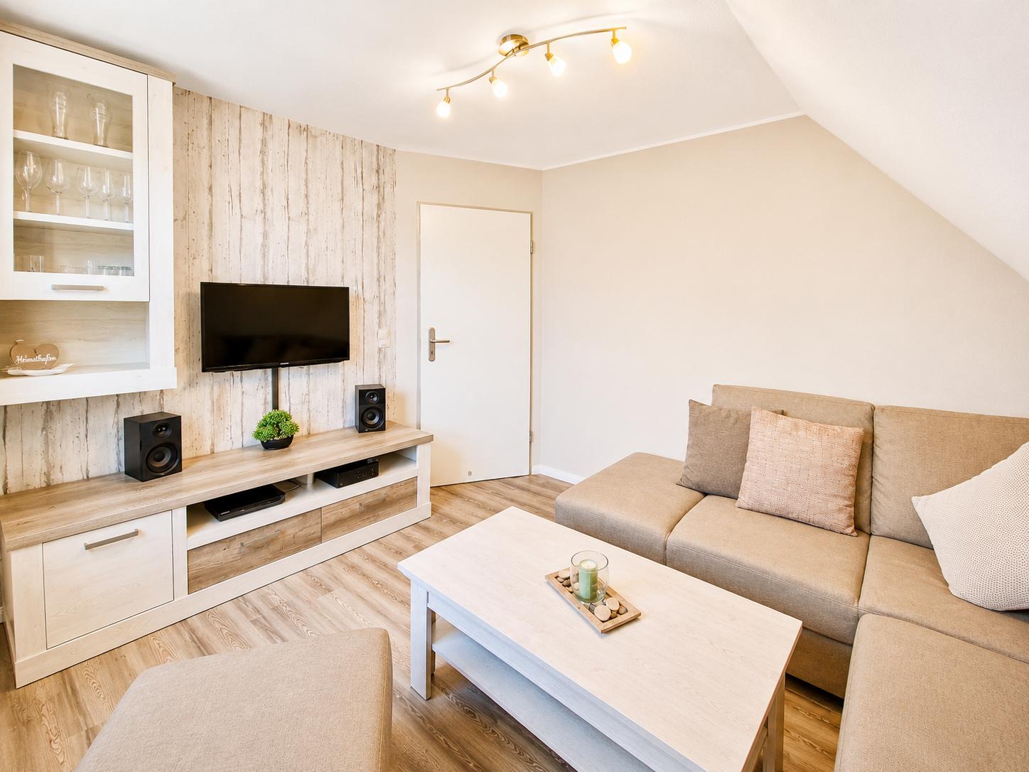 Wohnzimmer mit Sofa, Fernseher und Holzmöbeln. Dachschrägen und Lichtstrahler sorgen für Raumhöhe.