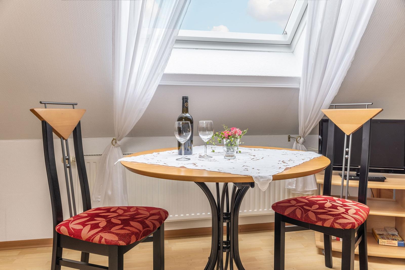 Runder Tisch mit zwei Stühlen unter Dachfenster. Tischdecke, Wein und Blumen.