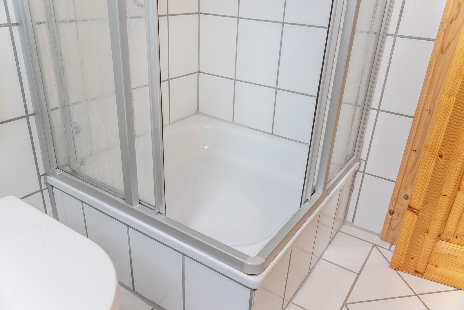 Dusche mit Glaswand und weißen Fliesen. Holztür und weißer Waschtisch sichtbar.