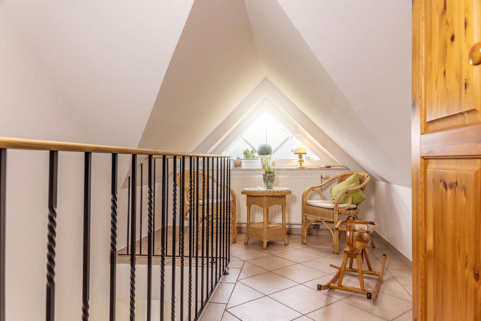 Dachgeschosszimmer mit Sitzgelegenheit und Fenster. Holzgeländer und Fliesenboden.
