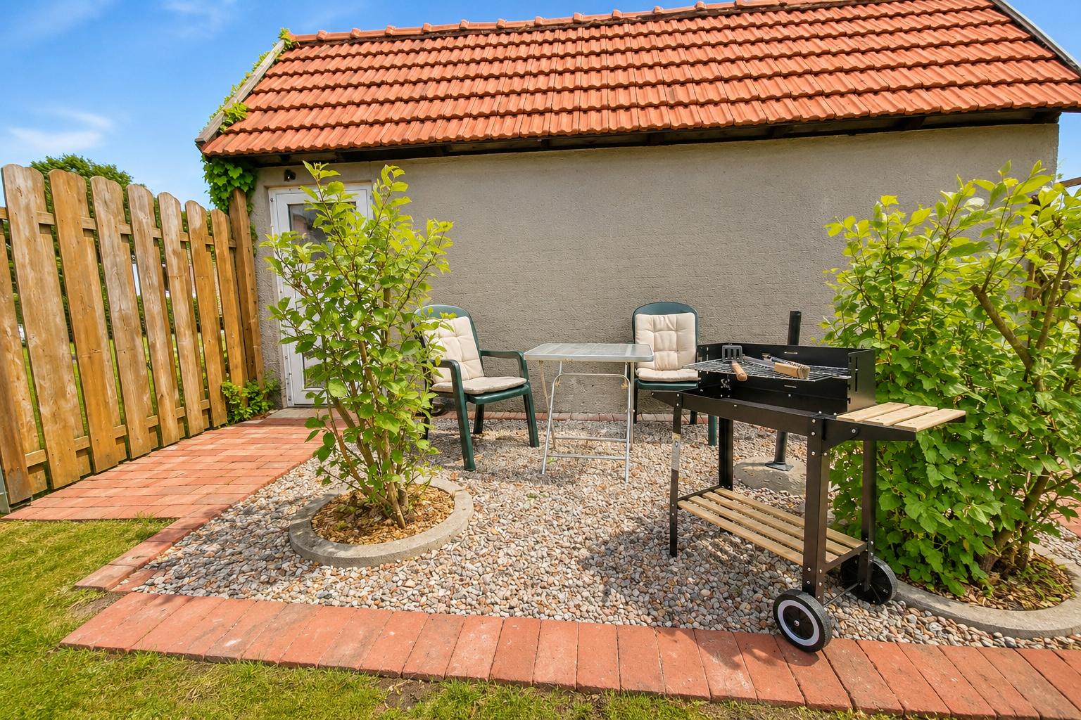 Hinterhof mit Grill, Tisch und Stühlen. Roter Ziegeldach, Holzzaun und Grasflächen.