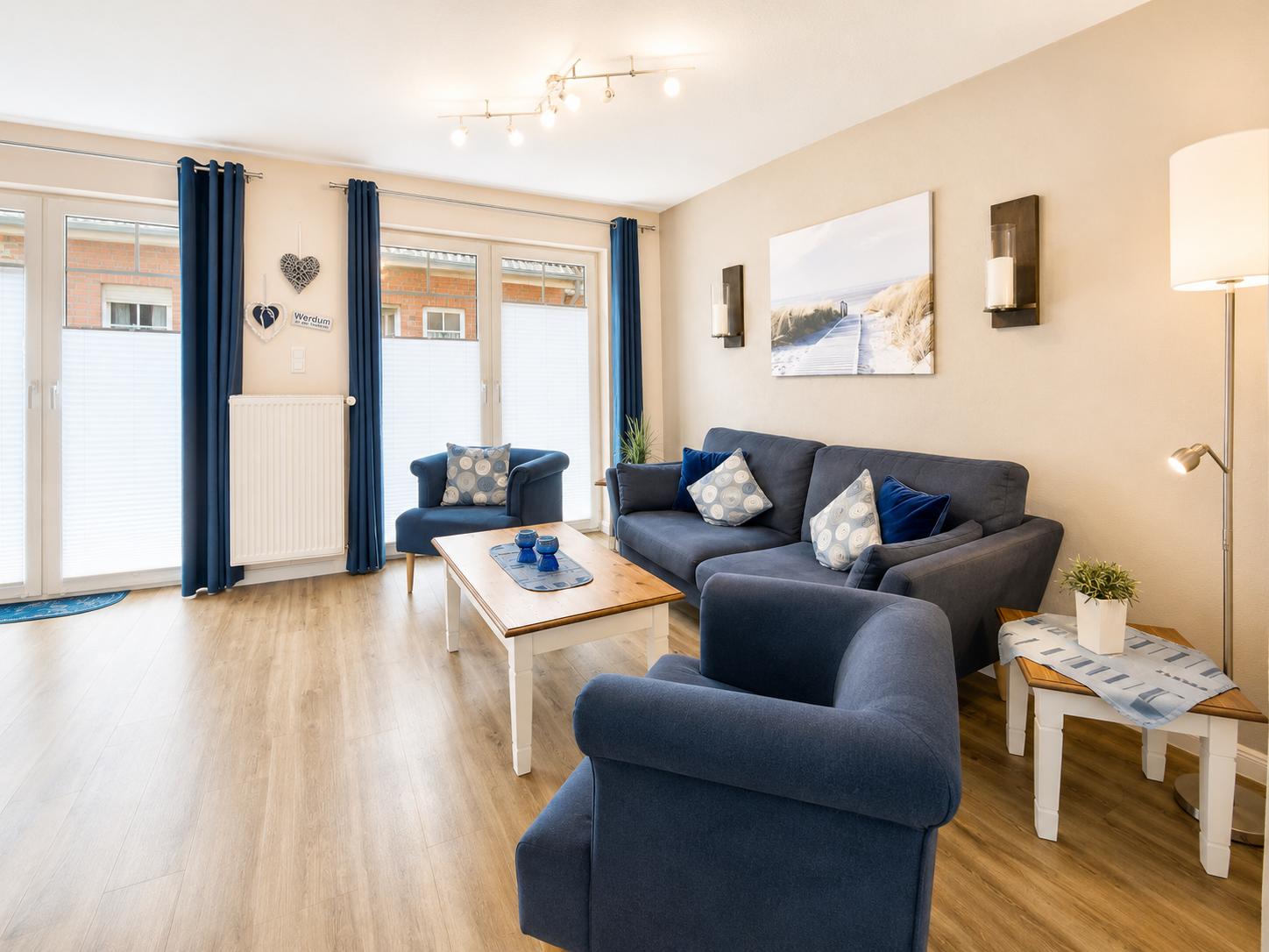 Wohnzimmer mit blauem Sofa, Sessel und Holztisch. Große Fenster mit blauen Vorhängen.