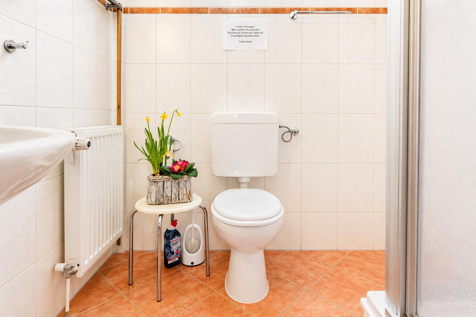 Badezimmer mit Toilette, kleinem Tisch und Blumen. Wand mit Hinweis und Heizkörper.