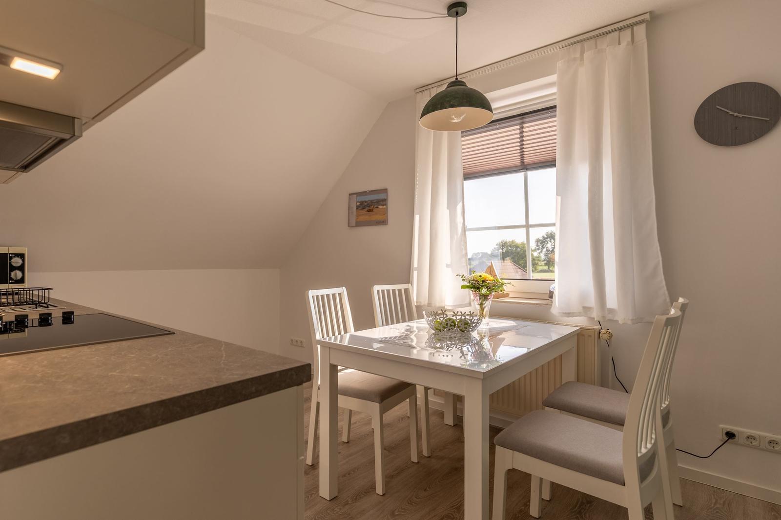 Essbereich mit weißem Tisch und Stühlen, Fenster mit Vorhängen und Küchenzeile.