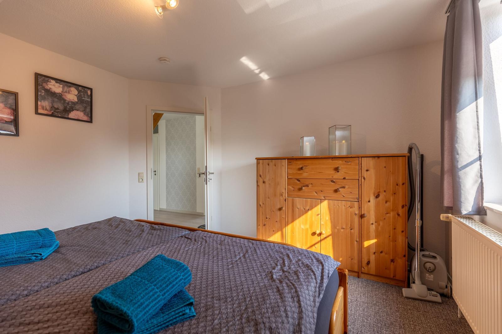 Schlafzimmer mit Bett, Holzschrank und Fenster. Auf dem Bett liegen gefaltete Handtücher.