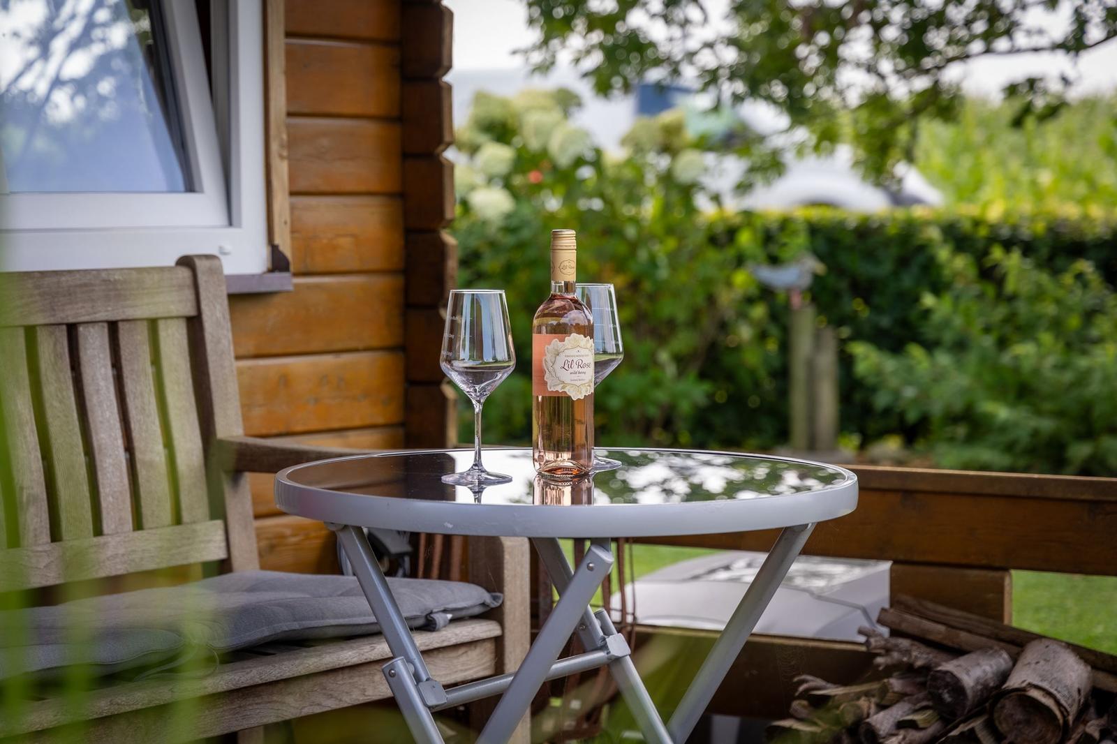Auf der Terrasse steht ein Tisch mit Weinflasche und zwei Gläsern. Hintergrund: Holzhaus und grüne Pflanzen.