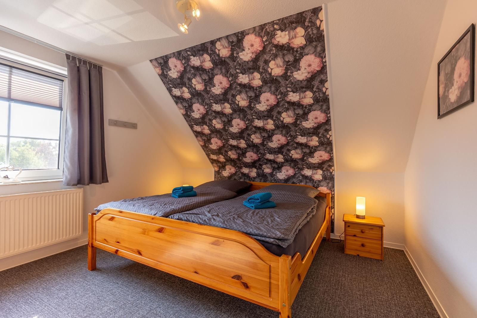 Schlafzimmer mit Holzbett, floraler Wandtapete und Nachttisch mit Lampe.