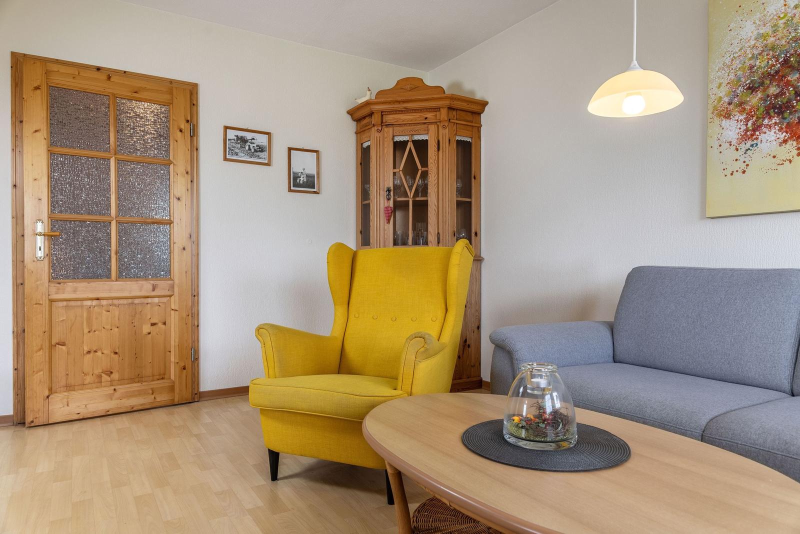 Wohnzimmer mit gelbem Sessel, grauem Sofa und Holztisch. Hintergrund: Holzschrank und Tür.