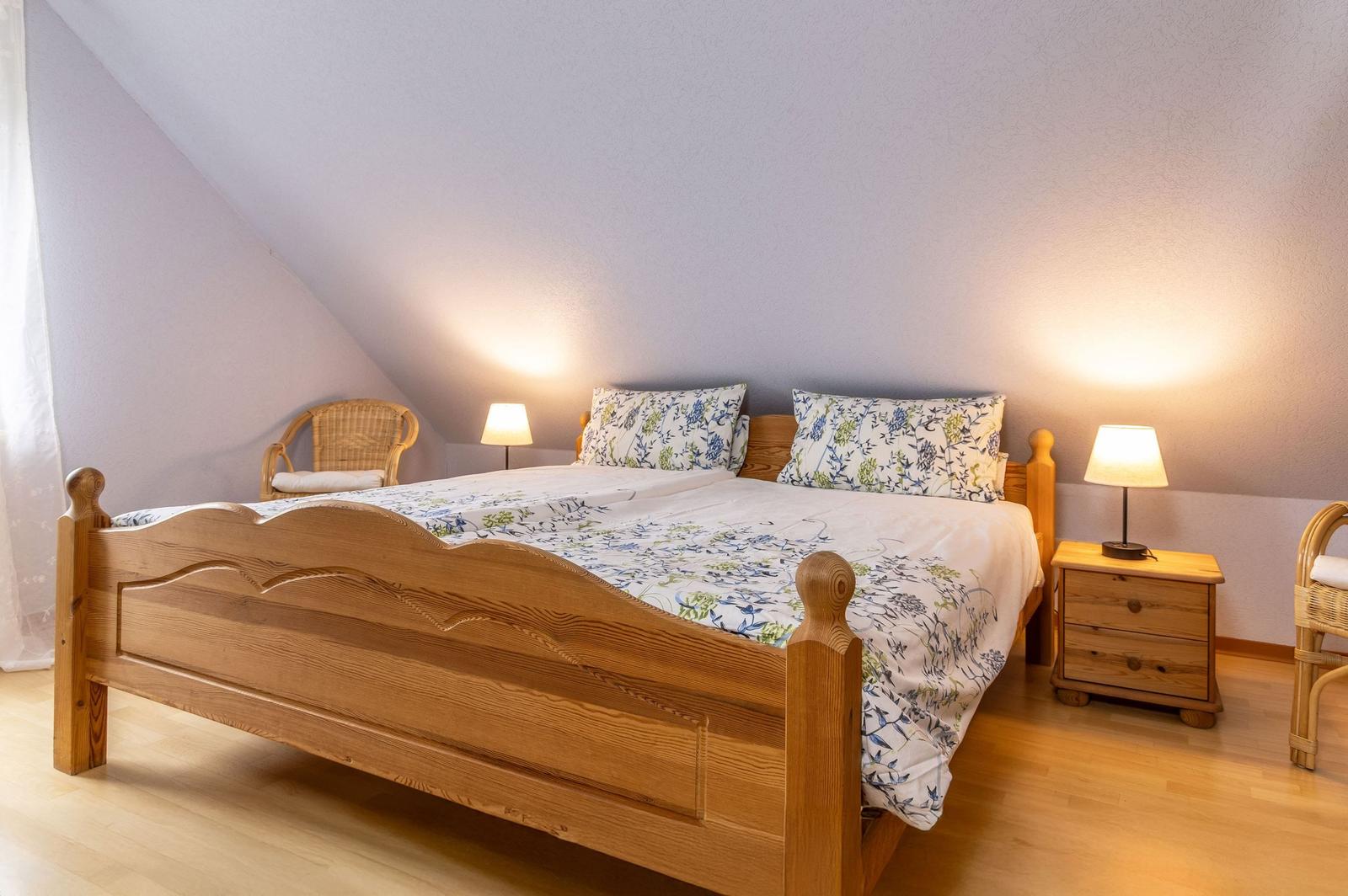Schlafzimmer mit Holzbett, Nachttisch und Wickerstühlen unter schrägen Wänden.