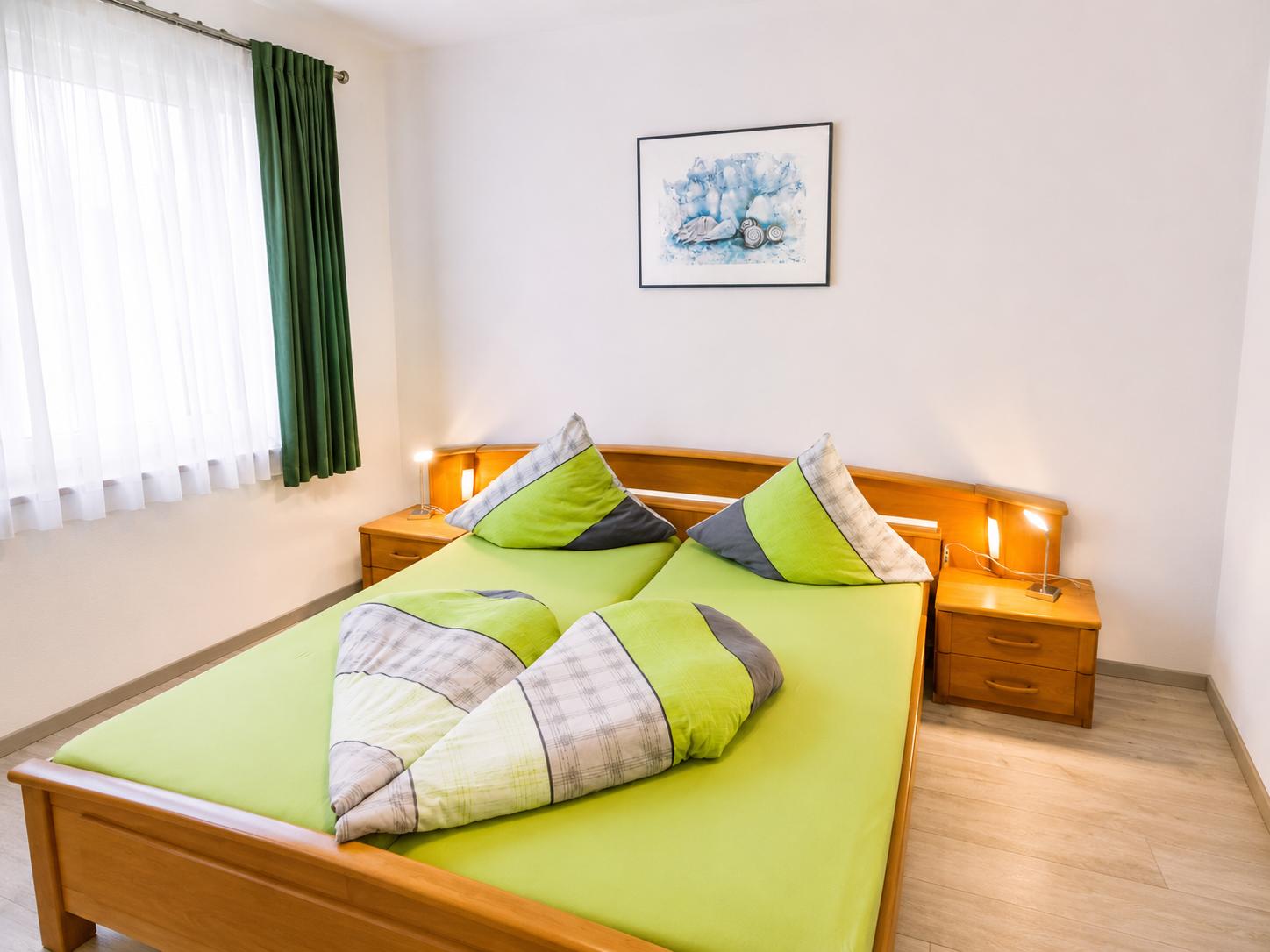 Schlafzimmer mit Doppelbett, grüner Bettwäsche, zwei Nachttischen und Fenster mit Vorhängen.