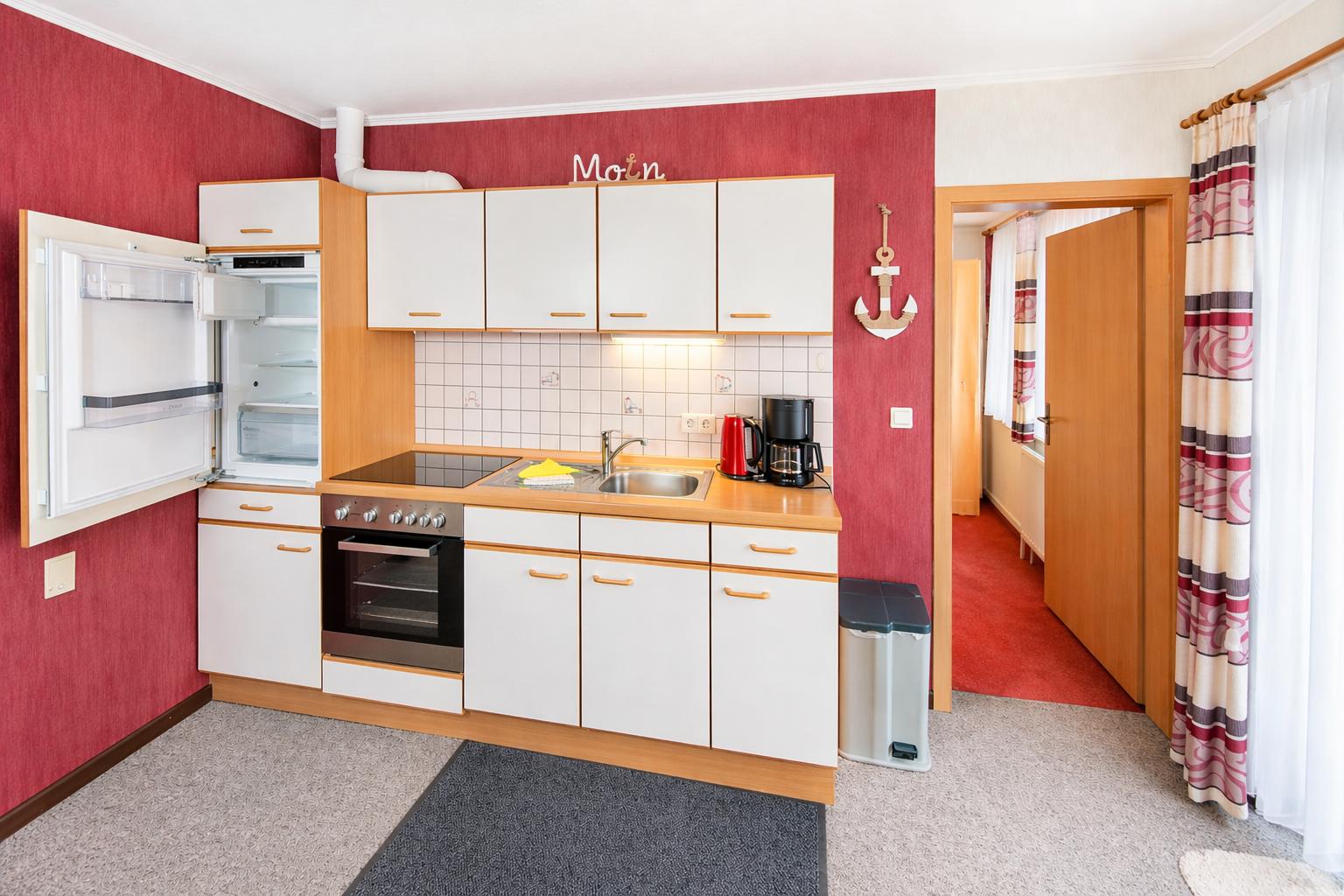 Küche mit weißen Schränken, Kühlschrank und Herd. Roter Wandverkleidung und Blick in den Flur.