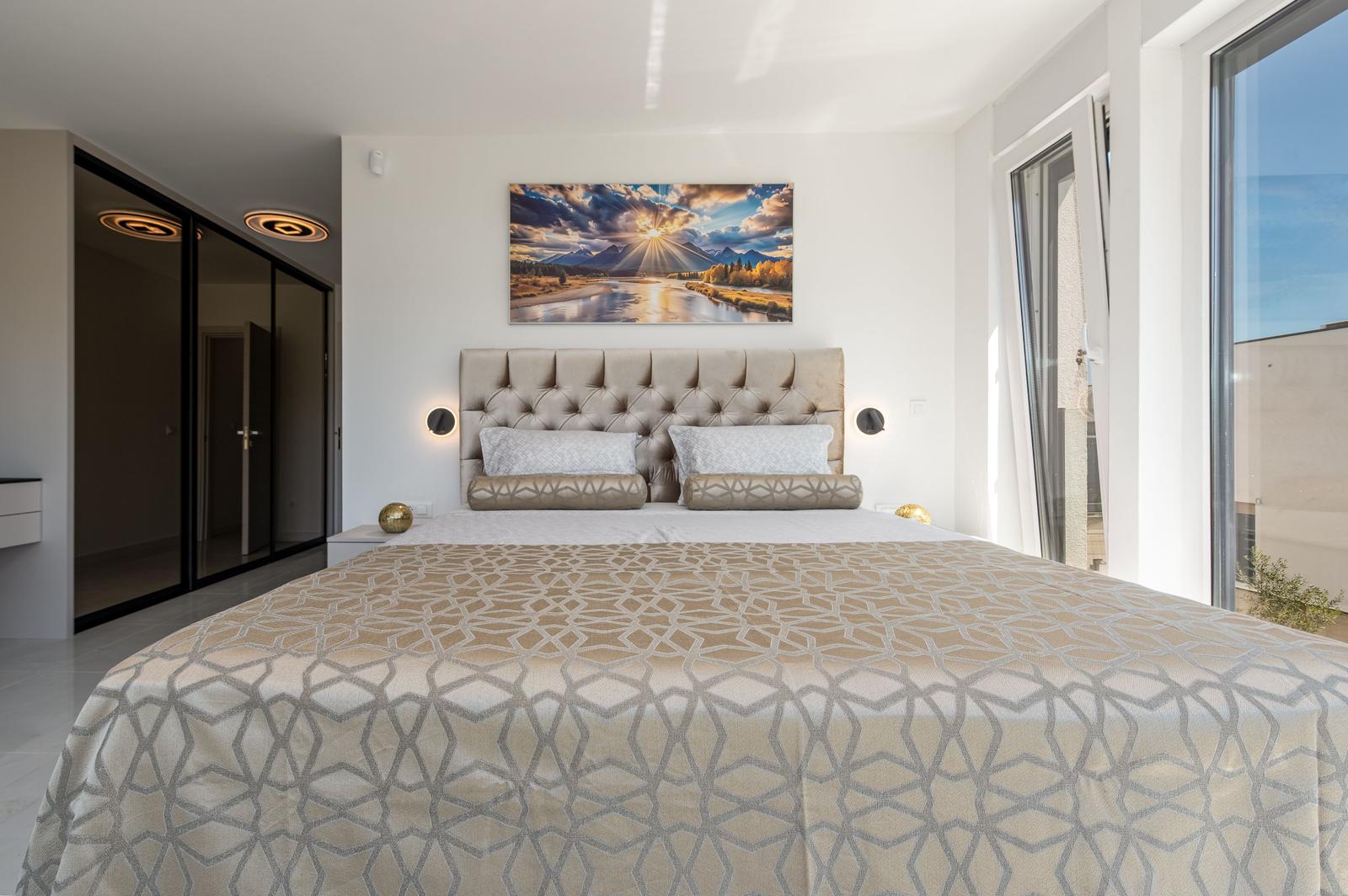 Schlafzimmer mit großem Bett, Wandbild und bodentiefen Fenstern.