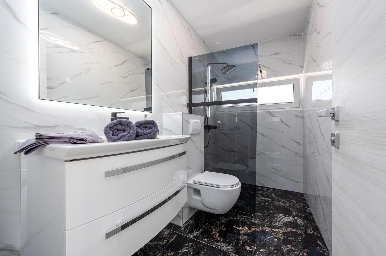 Badezimmer mit Waschbecken, Dusche und Toilette. Marmorwände und dunkler Boden.