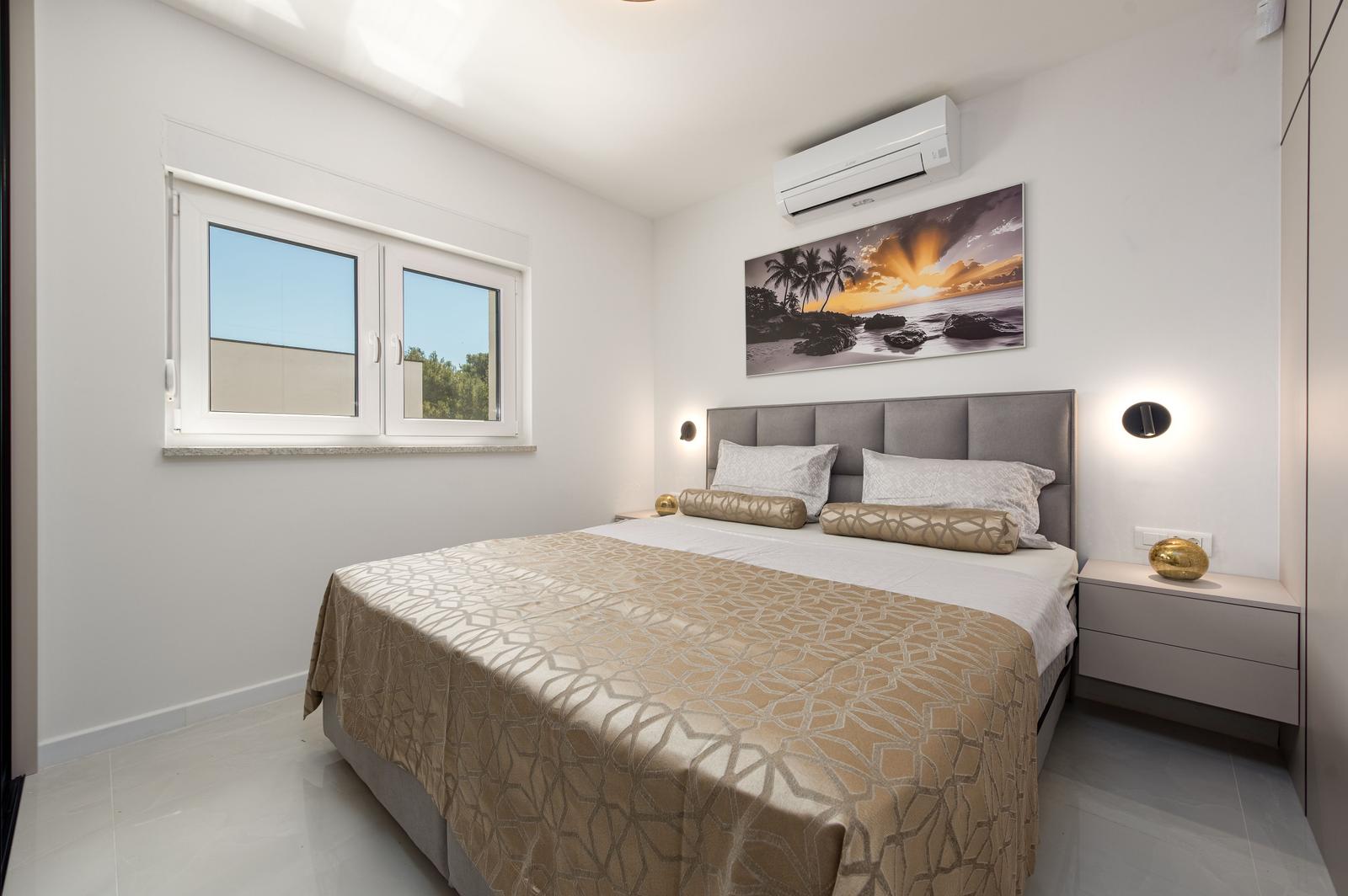 Schlafzimmer mit Bett, Nachttisch und Fenster. Wandbild und Klimaanlage sind sichtbar.