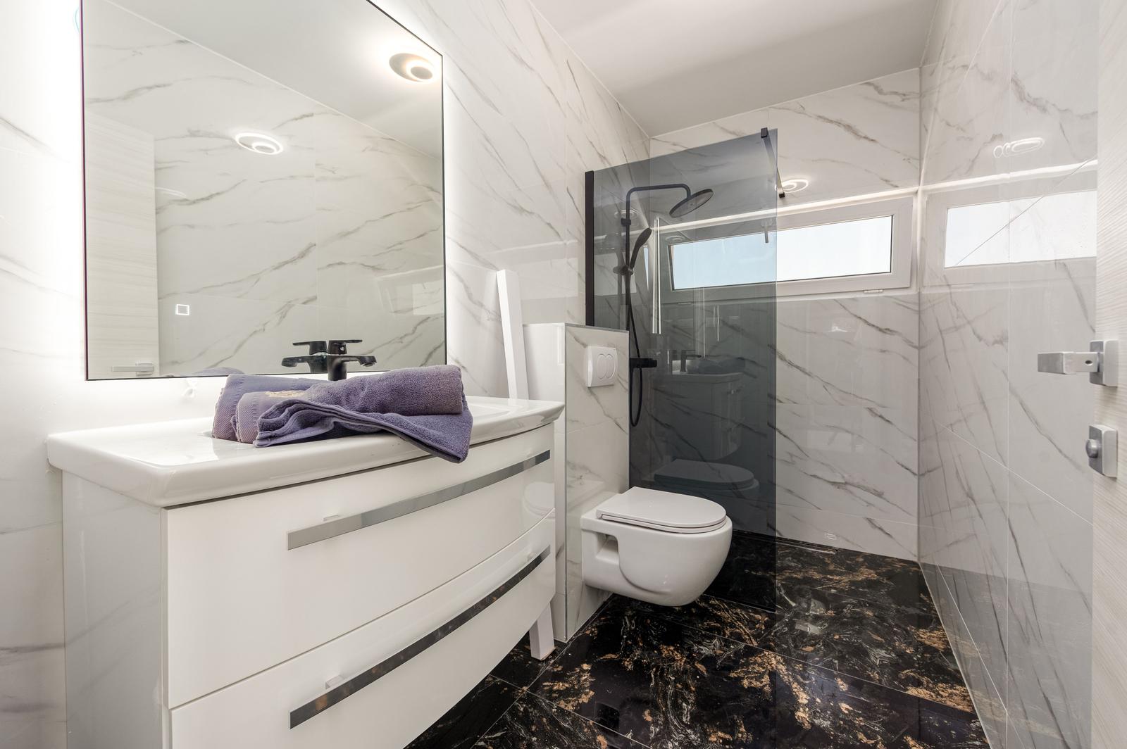 Badezimmer mit Dusche, Waschbecken und Toilette. Marmorwände und dunkler Boden.