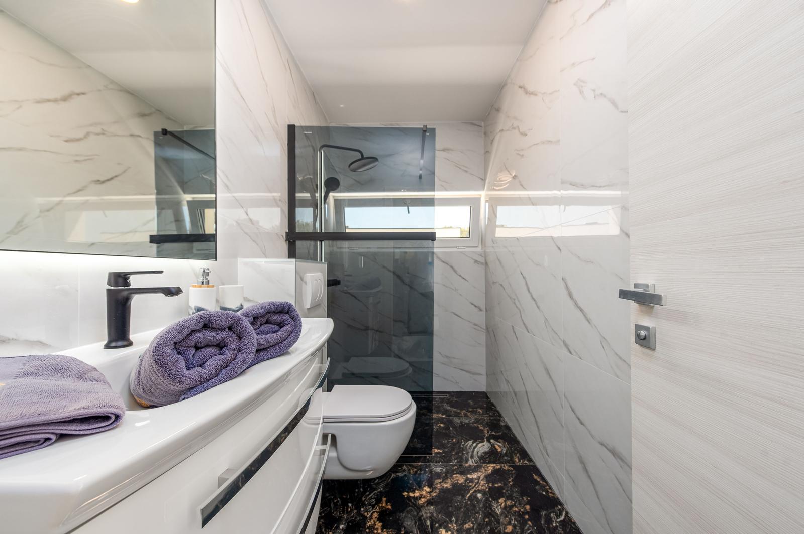 Badezimmer mit Dusche, Waschbecken und Toilette. Marmorwände und dunkler Boden.