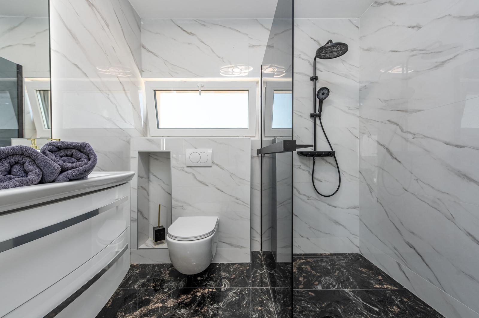Badezimmer mit Dusche, Toilette und Waschbecken. Weiße Marmorwände, schwarzer Boden und Fenster.